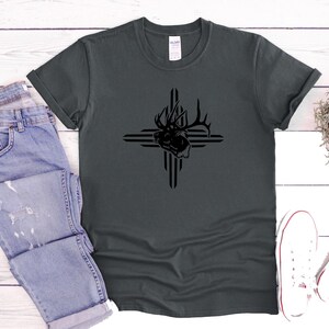 May include: Dark gray t-shirt featuring a black graphic of an elk head within a cross design. The shirt is paired with light blue denim jeans and white canvas sneakers. The t-shirt is made of a soft material.