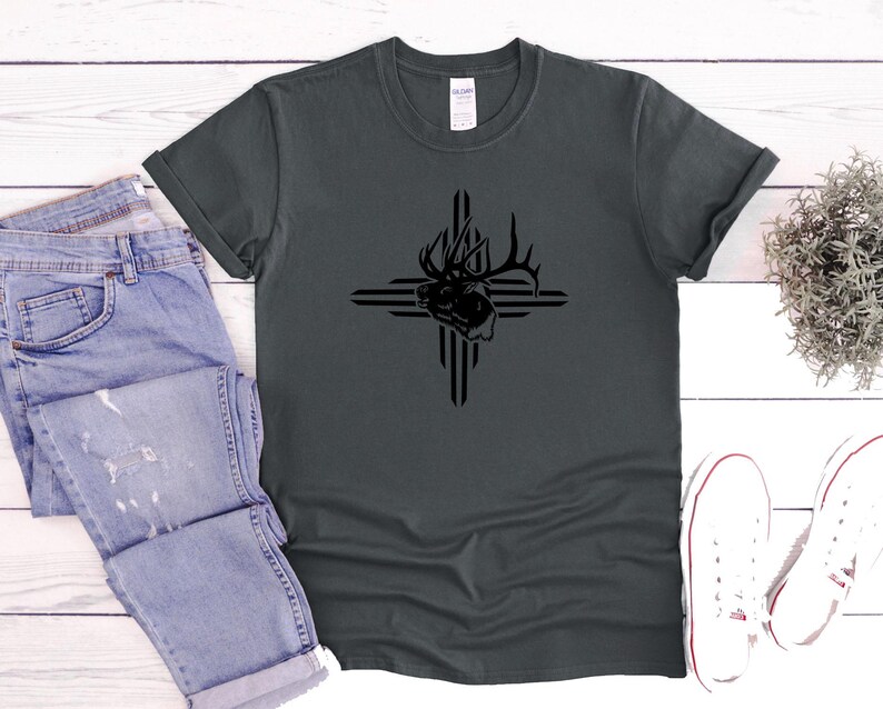 May include: Dark gray t-shirt featuring a black graphic of an elk head within a cross design. The shirt is paired with light blue denim jeans and white canvas sneakers. The t-shirt is made of a soft material.