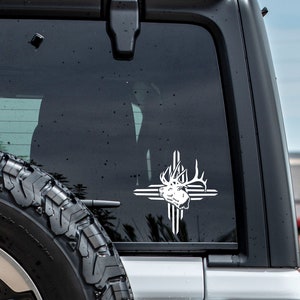 May include: White vinyl decal of a deer head with antlers, set within a cross design. The decal is affixed to the rear window of a black SUV, with a spare tire visible. The design is a Southwestern style.