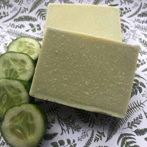 May include: Two rectangular bars of green soap with a textured surface, next to cucumber slices. The soap and cucumbers are on a white background with a green fern pattern. The soap is a muted green colour.
