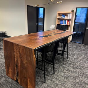 May include: A long, dark wood table with a black epoxy river running through the centre. The table is surrounded by four black bar stools. The table is in a modern office setting.