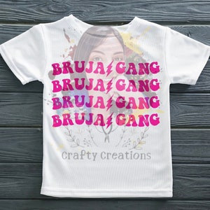 Könnte beinhalten: Weißes T-Shirt mit einem rosa-weißen Aquarelldesign, das das Gesicht einer Frau und den Text "BRUJA GANG" viermal wiederholt. Der Text ist in einem Retro-Schriftart-Stil. Der untere Teil des Shirts trägt den Text "Crafty Creations".