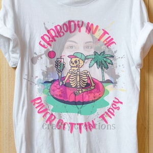 May include: White t-shirt with a pink graphic design featuring a skeleton in a pink inner tube floating in a turquoise river. The skeleton is wearing a green baseball cap and holding a green drink. The text "Everybody in the river gettin' tipsy" is written in pink around the graphic.