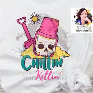 以下が含まれることがあります： ピンクのバケツに頭蓋骨、シャベル、そして「Chillin' Killin'」というテキストが描かれたカラフルなグラフィックがプリントされた白いTシャツです。