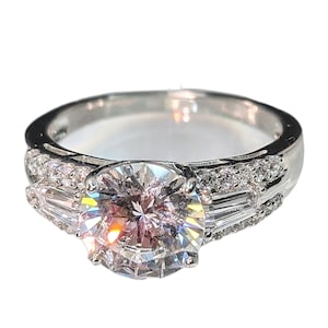 May include: A silver-toned ring featuring a large, round, faceted center stone. The band is accented with smaller, round, pavé-set stones and two baguette-cut stones flanking the center, creating a multi-faceted, reflective appearance.