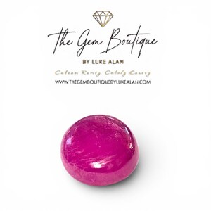 May include: A vibrant, polished, fuchsia-colored gemstone. The Gem Boutique logo is at the top, with the text "The Gem Boutique by Luke Alan" and website address. The gemstone is round and sits on a white background.