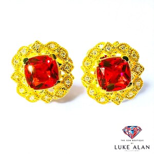Puede incluir: Un par de pendientes dorados. Cada pendiente presenta una gran gema roja cuadrada en el centro, rodeada por un detallado diseño de filigrana dorada con pequeñas piedras claras. Los pendientes están sobre un fondo blanco.