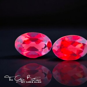 May include: Two oval-shaped red gemstones with a slight purple hue, set against a black background. The gemstones are smooth and polished, reflecting light.