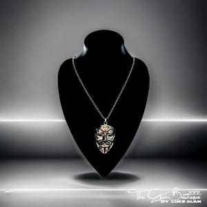 May include: A silver chain necklace with a silver pendant featuring a stylized face mask. The mask has a black and white design with red accents.