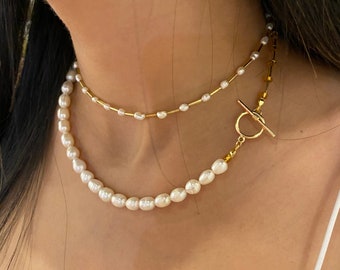 May include: A layered necklace with a delicate gold chain and a strand of white pearls. The necklace features a gold toggle clasp.