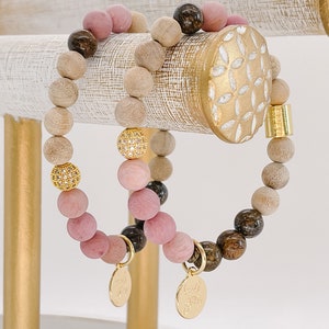 May include: Two wooden beaded bracelets with gold accents. One bracelet has pink and brown beads, the other has natural wood beads. Both bracelets have a gold charm with the words "Good Vibes" on them.