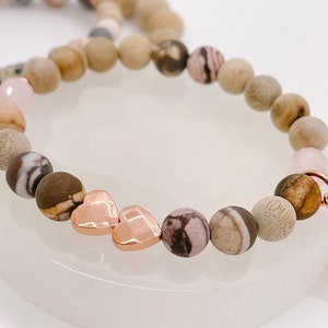 May include: Beaded bracelets with natural stone beads in brown, beige, and pink hues. The bracelets have rose gold heart charms and a round charm with a logo.