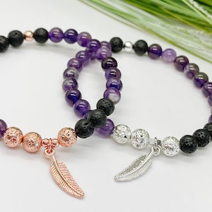May include: Two bracelets featuring amethyst and lava rock beads. One bracelet has a rose gold feather charm, the other has a silver feather charm.