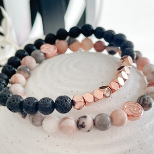 May include: Two bracelets, one with black lava rock beads and the other with pink and grey beads. Both bracelets have rose gold accents.