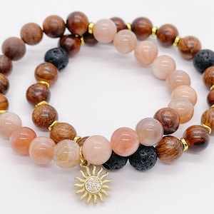 May include: A double-strand bracelet featuring brown wood beads, black lava beads, and pink sunstone beads. A gold sun charm dangles from the bracelet.