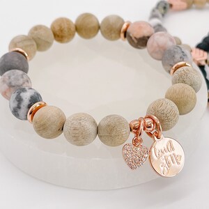 May include: A bracelet with wooden and speckled stone beads, with rose gold-coloured spacers. The bracelet has rose gold charms, a heart and a round charm with text. Other bracelets are visible in the background.