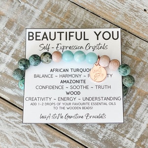 May include: A turquoise, green, and brown beaded bracelet with a wooden charm that says "Love you" in gold lettering. The bracelet is on a white card with the text "Beautiful You Self-Expression Crystals" and a list of the crystals and their meanings.