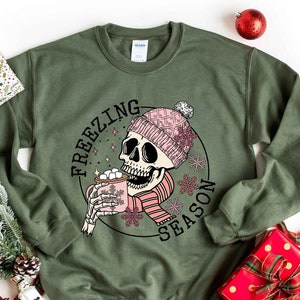 May include: Green sweatshirt with a graphic of a skeleton wearing a pink hat and scarf, holding a mug of hot chocolate. The text "Freezing Season" is written around the graphic.