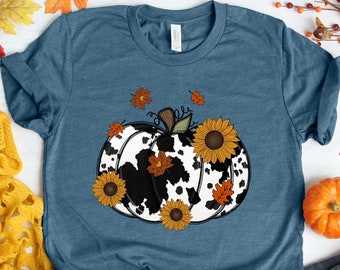 May include: A teal blue t-shirt with a white and black cow print pumpkin design. The pumpkin is surrounded by orange and brown leaves and yellow sunflowers.