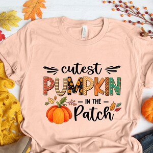 May include: A peach-colored t-shirt with the text "cutest pumpkin in the patch" in black and colorful letters. The text is surrounded by a small pumpkin graphic and fall leaves.