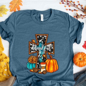 May include: Teal blue t-shirt with a white and black cross design. The cross has the word "thankful" written on it. The cross is surrounded by fall-themed elements, including pumpkins, gourds, and pinecones.