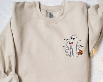 Könnte beinhalten: Ein beiger Pullover mit einem Geist, der ein Glas Wein und einen Kürbis hält. Der Geist ist schwarz umrandet und hat ein kleines Lächeln. Um den Geist herum fliegen Fledermäuse.