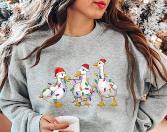 Op de afbeelding: Een grijs sweatshirt met een kerstthema ontwerp met drie witte eenden die kerstmanmutsen dragen en zijn gewikkeld in kleurrijke kerstverlichting.