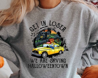 May include: Gray sweatshirt with a graphic print of a yellow taxi cab with a skeleton driver. The text on the shirt reads "Get in Loser, We are saving Halloweentown."