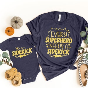 May include: A navy blue t-shirt with gold glitter text that reads "Every Superhero Needs A Sidekick". A matching navy blue bodysuit with gold glitter text that reads "Sidekick" is next to it.