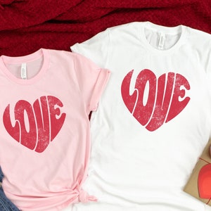 May include: Two t-shirts featuring a red heart-shaped design that says "LOVE" in a retro font. The t-shirts are one pink and one white.