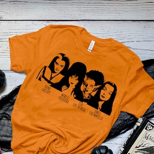 May include: Orange t-shirt with a black silhouette of the Addams Family characters. The text reads "Think Like Lily", "Work It Like Elvira", "Love Like The  Addams", "Dance Like Morticia".