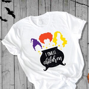 May include: White t-shirt with a black cauldron and three witches with the text "I smell children" printed on it. The witches have purple, orange, and yellow hair.