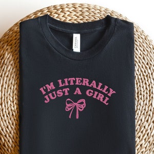 May include: Black t-shirt with the text "I'M LITERALLY JUST A GIRL" in pink, arched above a pink bow graphic. The shirt is folded and placed on a round, woven, light brown surface.