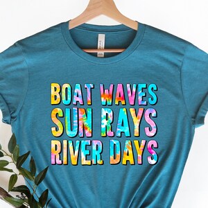 May include: Teal t-shirt with the words "BOAT WAVES SUN RAYS RIVER DAYS" in a colorful tie-dye design. The shirt is displayed on a wooden hanger against a white background. A sprig of greenery is placed near the bottom.