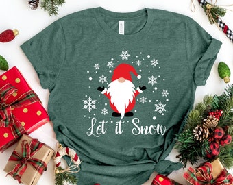 Könnte beinhalten: Ein grünes T-Shirt mit einer weißen Grafik eines Gnoms mit roter Mütze und Bart, umgeben von Schneeflocken. Der Text "Let it Snow" ist unter dem Gnom gedruckt. Das T-Shirt ist umgeben von Weihnachtsdekorationen, darunter verpackte Geschenke, Zuckerstangen und Kiefernäste.