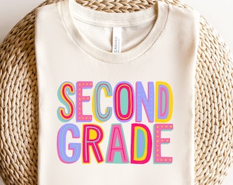 Könnte beinhalten: Ein weißes T-Shirt mit den Worten "Second Grade" in bunten, verspielten Buchstaben. Die Buchstaben sind mit rosa und gelben Punkten verziert.