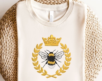 May include: A white t-shirt with a gold embroidered bee and crown design. The bee is surrounded by a wreath of gold leaves.