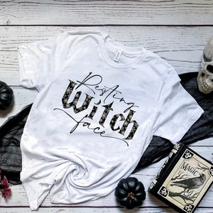 May include: White t-shirt with the text "Resting Witch Face" in black script with a black and white pattern. The shirt is surrounded by Halloween decorations, including a black skull, black ravens, and black pumpkins.