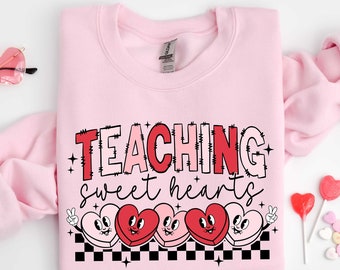 May include: Pink sweatshirt with the text "TEACHING sweet hearts" in red and black. The text is surrounded by a row of five cartoon hearts with faces.