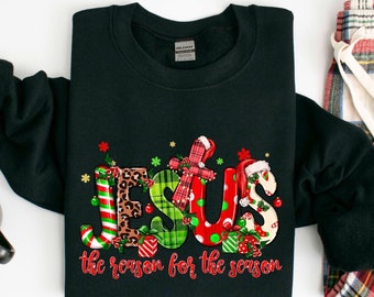 May include: Black sweatshirt with a Christmas-themed design featuring the word "JESUS" in colorful letters with a festive pattern. The text below reads "the reason for the season".