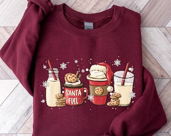 May include: A maroon sweatshirt with a Christmas-themed design featuring a Santa hat, a cup of coffee, a glass of milk, and cookies. The text "Santa Fuel" is printed on the sweatshirt.