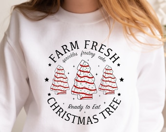 Op de afbeelding: Witte trui met een zwart grafisch ontwerp van drie kerstboom met rode en groene frosting, sprinkles en de tekst "Farm Fresh Christmas Tree" met de woorden "sprinkles frosting cake" en "Ready to Eat" in een kleiner lettertype.