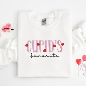 May include: White sweatshirt with the text "CUPID'S favorite" in pink lettering with red hearts.