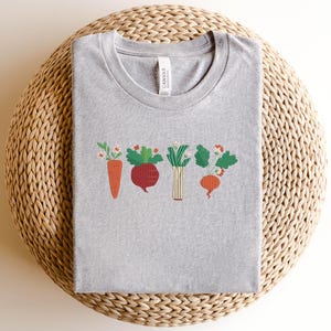 May include: Grey t-shirt featuring a graphic of a carrot, beetroot, onion, and turnip with green leaves and flowers. The shirt has a crew neck and short sleeves.