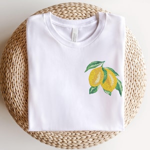 May include: White t-shirt with an embroidered lemon design. The lemons are yellow with green leaves.