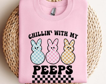 May include: Pink sweatshirt with the text "Chillin' with my Peeps" in black. Three cartoon peeps are shown in blue, pink, and yellow with white checkered patterns.