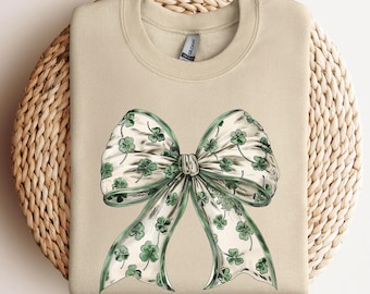 May include: A beige sweatshirt with a green shamrock bow design. The bow is made of white ribbon with green shamrocks printed on it.