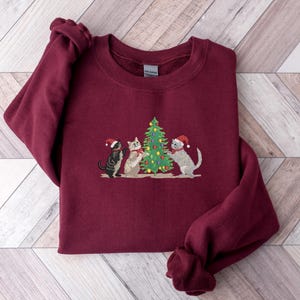 K&ouml;nnte beinhalten: Ein weinrotes Sweatshirt mit einem gestickten Design von drei Katzen mit Weihnachtsm&uuml;tzen, die sich um einen geschm&uuml;ckten Weihnachtsbaum versammelt haben. Die Katzen sind in den Farbt&ouml;nen Schwarz, Grau und Wei&szlig; gehalten. Das Sweatshirt hat einen Rundhalsausschnitt und lange &Auml;rmel.