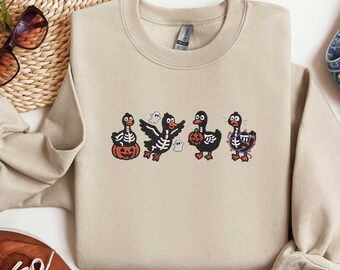 May include: Beige crewneck sweatshirt featuring a Halloween-themed design. The design includes four cartoon ducks dressed in Halloween costumes, including a pumpkin, skeleton, and witch. The sweatshirt is paired with blue jeans and brown shoes.