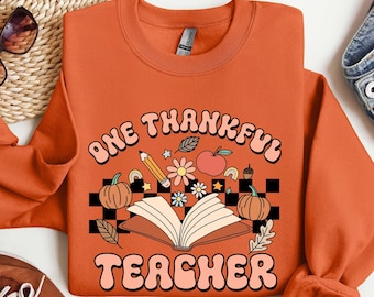 May include: Orange sweatshirt with the text "One Thankful Teacher" in a retro font. The design includes a book, pencils, pumpkins, and fall leaves.